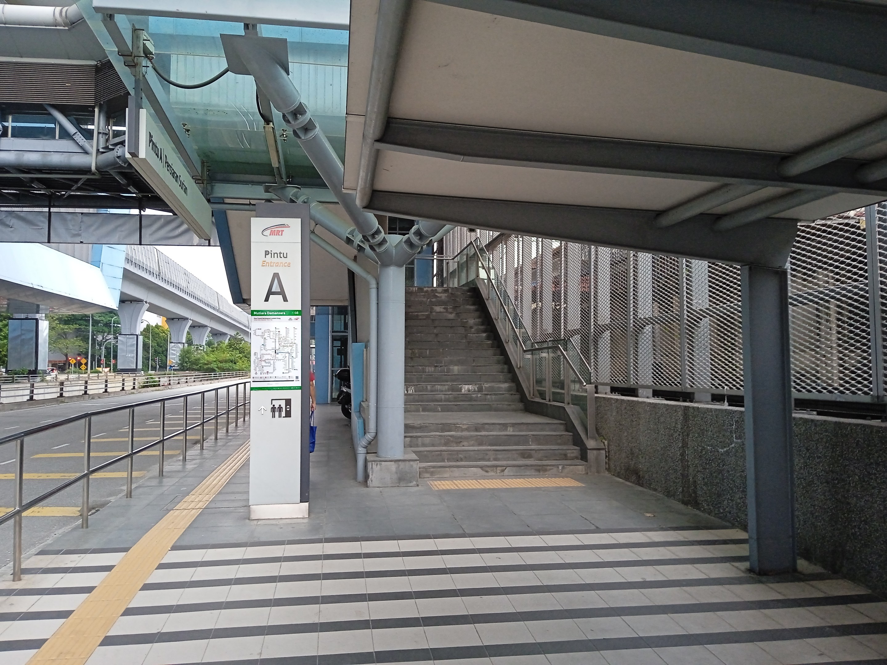 Ein Bahnsteig mit Treppe, eine Straße mit Geländer auf der linken Seite und ein Hintergrund mit Bäumen, Pfählen, Gebäuden und einer Brücke, mit Rohren und einer Tafel am oberen Ende des Bahnsteigs.