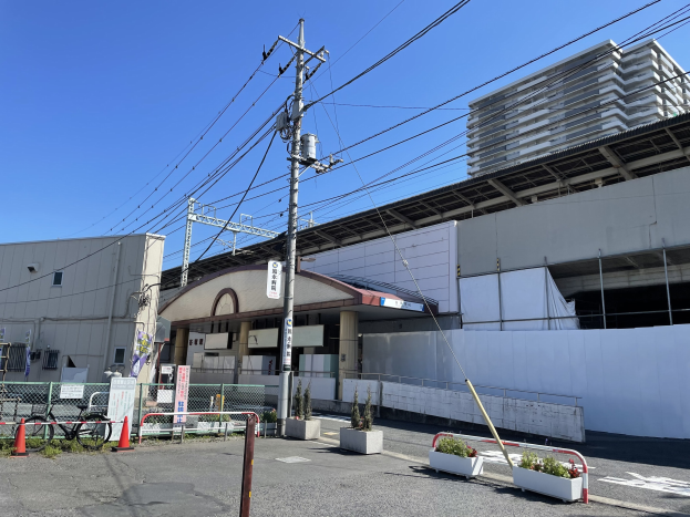 Großes Gebäude mit zahlreichen Stromleitungen davor, umgeben von städtischer Infrastruktur einschließlich Strommasten, Kabeln, Fahrzeugen, Absperrpollern, Verkehrskegeln, Topfpflanzen, Namensschildern, Geländern und einem klaren blauen Himmel.