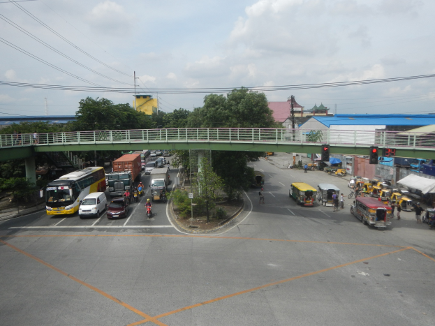 Eine belebte Straße mit Autos, Bussen, Fußgängern auf einem Gehweg, eine Brücke mit Geländern, Verkehrsampeln, Bäume, Gebäude, Strommasten mit Drähten und einen bewölkten Himmel.