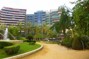 Ein Park mit einem zentralen Brunnen, umgeben von Grünflächen einschließlich Gras, Pflanzen und Bäumen, mit Bänken, Laternenmasten und Fahrzeugen auf einer nahen Straße sowie hohen Gebäuden und einem klaren blauen Himmel im Hintergrund.