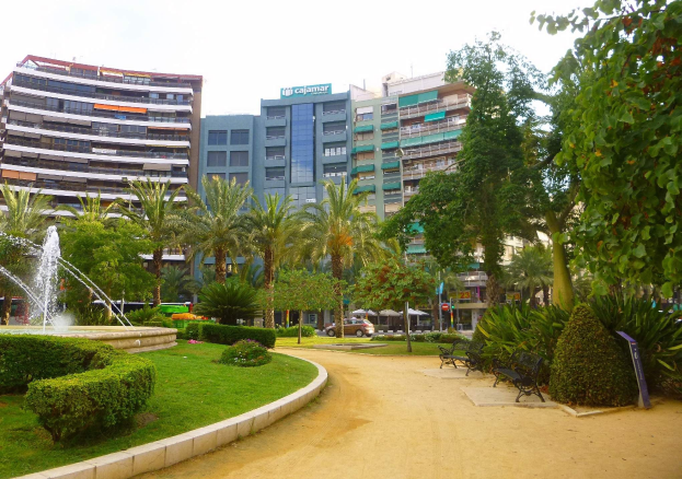 Ein Park mit einem zentralen Brunnen, umgeben von Grünflächen einschließlich Gras, Pflanzen und Bäumen, mit Bänken, Laternenmasten und Fahrzeugen auf einer nahen Straße sowie hohen Gebäuden und einem klaren blauen Himmel im Hintergrund.