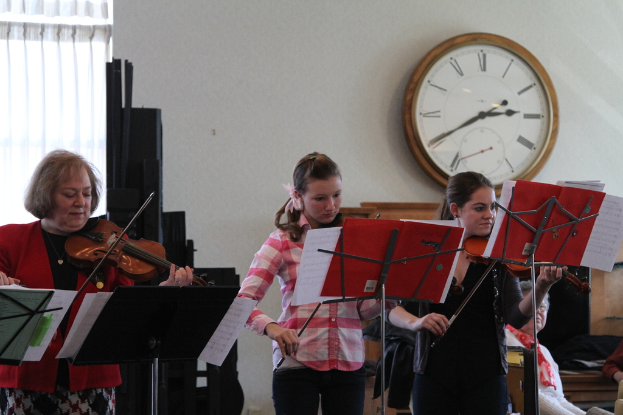 Drei Frauen auf der B√ľhne mit Musikst√§nden, die Geige spielen, w√§hrend eine Gruppe sitzender Frauen im Vordergrund zuh√∂rt; eine Uhr h√§ngt an der wei√üen Wand im Hintergrund, mit einem wei√üen Vorhang in der linken Ecke.