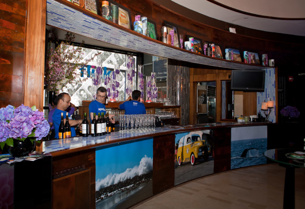 Ein Restaurant-Innenraum mit drei Personen, die hinter einem Tisch mit Gläsern, Flaschen, Aufklebern, Postern, einer Zimmerpflanze und Lampen arbeiten, mit einem Fernseher und Regalen im Hintergrund.