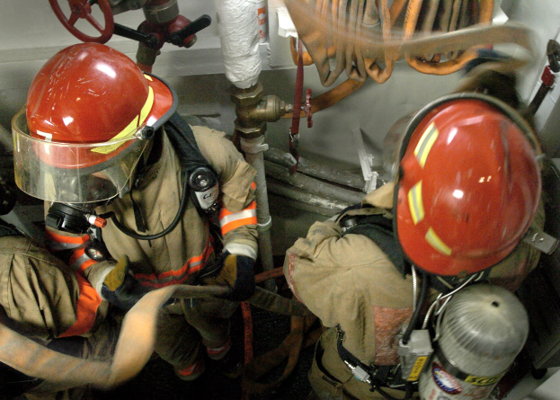 Feuerwehrleute in Schutzausrüstung arbeiten an einem Feuerhydranten in der Nähe von Rohren, Ventilen und einer Wand, mit Text am unteren Bildrand.