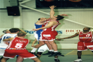 Männer beim Basketballspielen auf einem Court mit einer Wandlautsprecher und Text im Hintergrund, ein Basketball schwebt über ihnen.