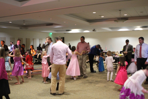 Eine Gruppe von Kindern und Erwachsenen, die um Tische mit Gegenständen darauf stehen, Stühle und von der Decke hängende Lampen.