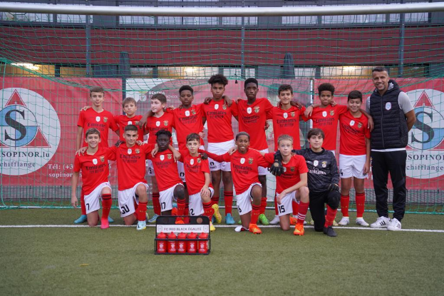 Eine Gruppe junger Männer in roten T-Shirts und schwarzen Shorts steht auf einem Fußballfeld neben einer Kiste roter Gegenstände, mit Schildern im Hintergrund, die "FC Red Black Eglaute - Champions League" lesen.