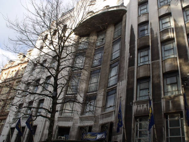 Gebäude mit Fenstern, eine Fahne und Flaggen an Stangen unten sowie ein Baum mit Zweigen.