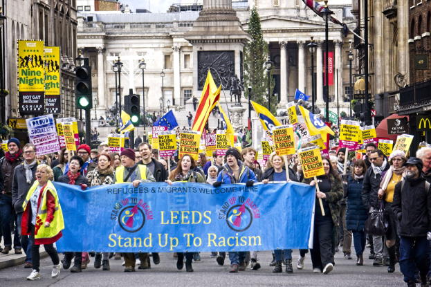 Gruppe von Menschen marschiert auf einer Stadtstraße mit einem 'Stand Up to Racism'-Schild, mit Gebäuden, Bäumen, Straßenlaternen, Ampeln und einer Statue im Hintergrund unter einem bewölkten Himmel.