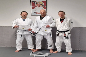 Drei Männer in weißen Judo-Uniformen stehen nebeneinander in einer Turnhalle, jeder hält eine Urkunde in der Hand, im Hintergrund hängt ein Rahmen an der Wand, der auf einen ersten Platz hindeutet.