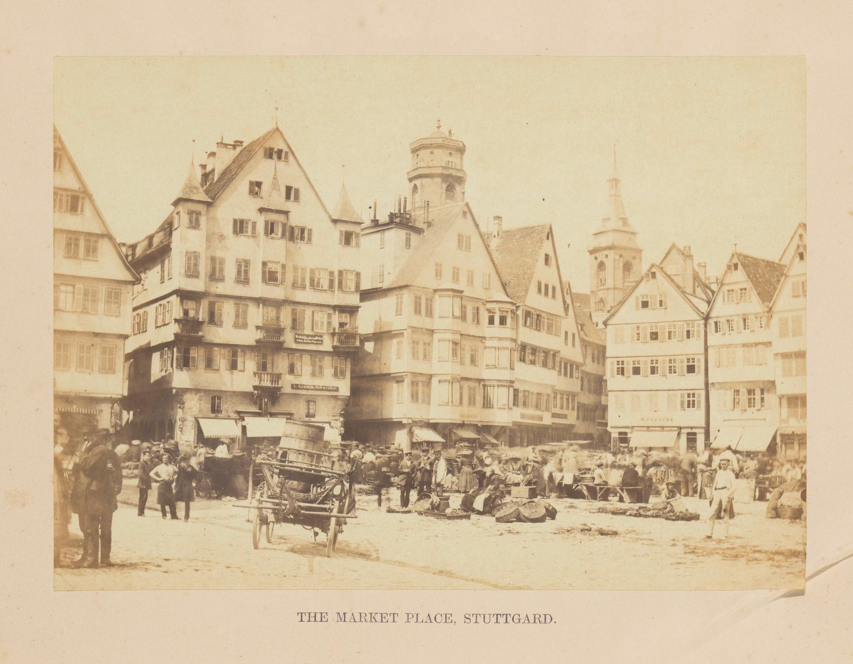 Schwarz-weißes Foto eines belebten Marktplatzes in Stuttgart, Deutschland, mit Gebäuden mit Fenstern, Menschen, Karren und diversen Gegenständen sowie Text unten.