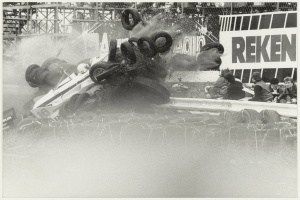 Ein Schwarz-Weiß-Foto eines verunglückten Rennwagens umgeben von Reifen, mit einer Gruppe von Menschen auf der rechten Seite und einer Tafel im Hintergrund.