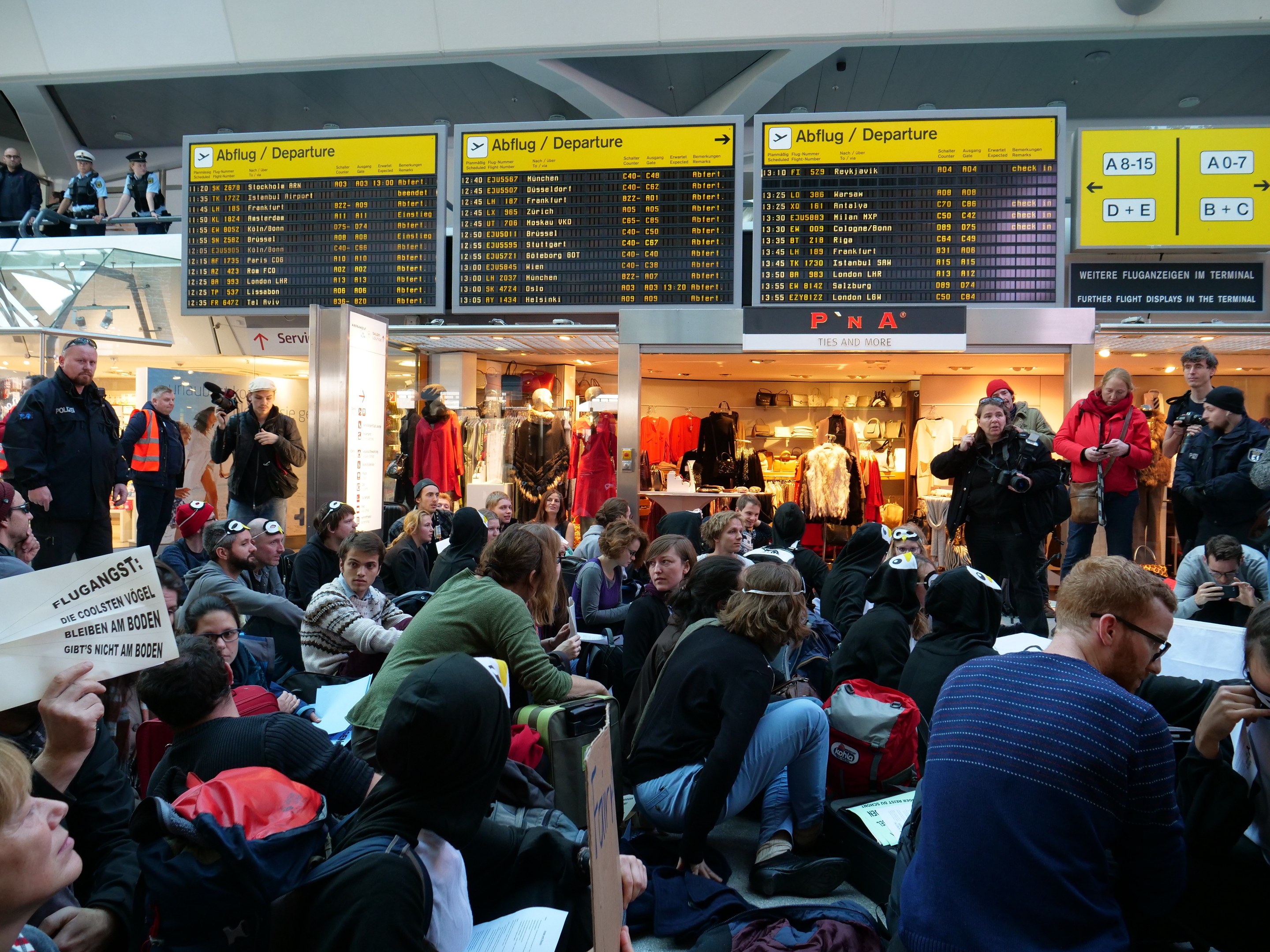Eine große Gruppe von Menschen sitzt und steht in einem Flughafen während einer Protestaktion, einige halten Taschen und Papiere, Schilder mit Text im Hintergrund, Schaufensterpuppen mit Kleidern und Deckenleuchten.