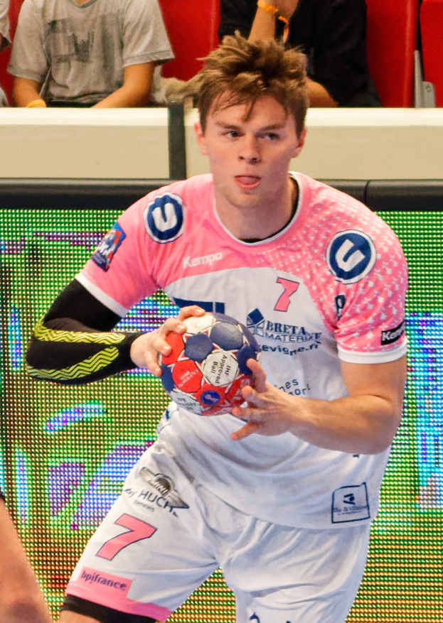 Ein Mann in einem pinken Hemd mit einem Fußball, daneben ist eine andere Person Hand zu sehen, während im Hintergrund Menschen auf Stühlen sitzen.