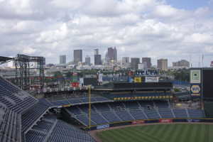 Baseballstadion mit Stadtpanorama im Hintergrund, gefüllt mit Stühlen, Pfählen und Brettern, auf Gras mit Bäumen und Gebäuden in der Ferne unter einem bewölkten Himmel.