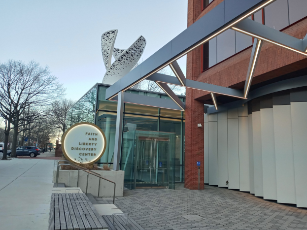 Eingang zum Faith and Liberty Discovery Center mit Glaswänden, einem Namensschild, einer Bank, einem Metallrahmen, Bäumen, Fahrzeugen, Pfosten und einem bewölkten Himmel.