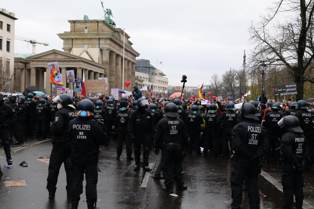 Eine große Gruppe von Polizisten steht vor einer Menge mit Schildern, Luftballons und Helmen, mit Gebäuden, Bäumen, einer Statue, einem Kran und einem bewölkten Himmel im Hintergrund.