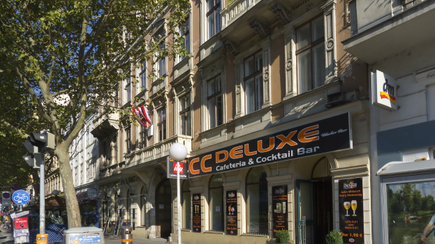 Eine belebte Stadtstraße mit verschiedenen Gebäuden, Fahrzeugen, Fußgängern und Schildern, darunter ein großes Gebäude mit der Aufschrift "CC Deluxe Cafe & Cocktail Bar" in der Mitte.