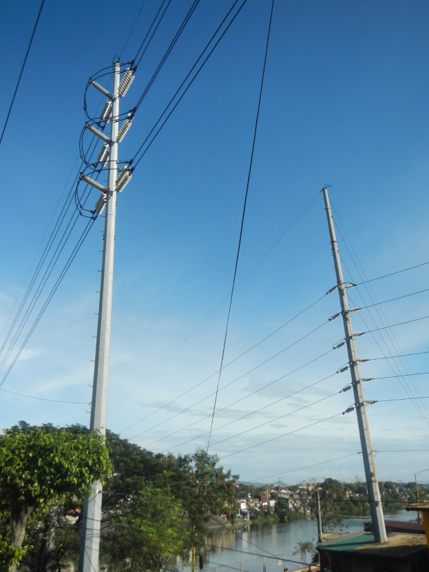 Leitung mit Strommasten und Drähten entlang einer Straße neben einem Gewässer, mit Bäumen und Gebäuden im Hintergrund unter einem klaren blauen Himmel.