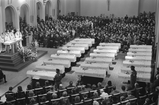 Eine große Gruppe von Menschen, die in einer Kirche sitzen, mit einigen, die vorne stehen, ein Podium mit einem Mikrofon auf der linken Seite und Blumensträuße und Lichter im Hintergrund, alles in Schwarz-Weiß.