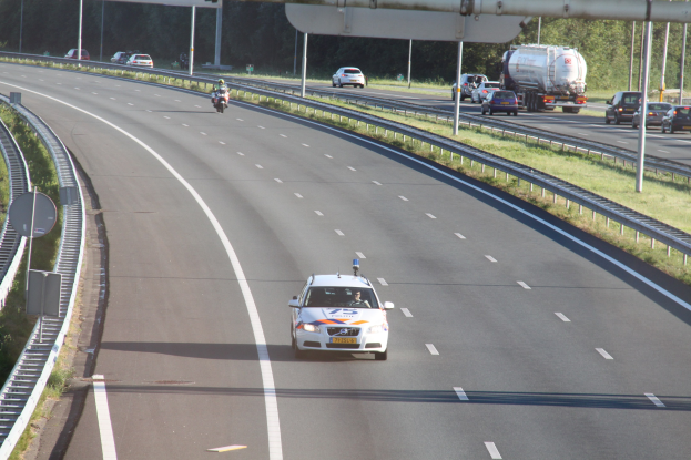 Ein Auto auf einer Autobahn mit einem stationären Polizeiauto am Straßenrand, andere Fahrzeuge und Bäume, Geländer, Pfosten und Planken im Hintergrund.