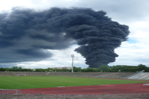 Ein Stadion mit einer großen schwarzen Rauchwolke, die aus seiner Spitze aufsteigt, umgeben von Gras, Stufen, Geländern, Bäumen, einem Lichtmast und einem bewölkten Himmel.