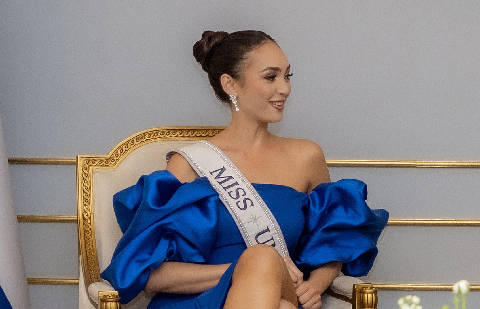 Frau in einem blauen Kleid mit einem "Miss Universe 2019"-Band, das auf einem Stuhl sitzend und von Blumen umgeben ist und lächelt.