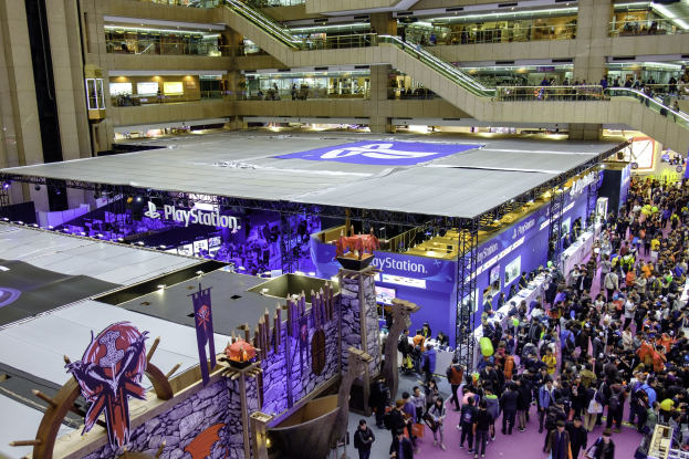Eine gro├če Menge Menschen in einem gro├čen Geb├Ąude mit Rolltreppen, Gel├Ąndern, beleuchteten Schildern, Bannern und anderen Convention-Ausstellungen, mit einem Wasserzeichen, das es als die weltweit gr├Â├čte Gaming-Convention ausweist.