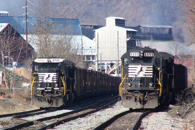 Zwei Züge auf parallelen Gleisen mit Bäumen, Strommast und Gebäuden im Hintergrund.