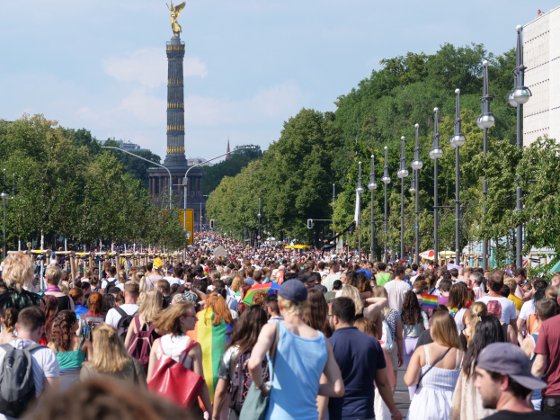 Eine große Menschenmenge geht eine Straße in Berlin, Deutschland, entlang und trägt Taschen mit Leuchtmasten am Weg, Bäumen und einem Turm mit einer Statue im Hintergrund, umgeben von Gebäuden unter einem bewölkten Himmel.