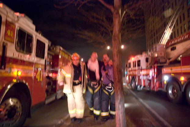 Drei Personen vor einer Feuerwache auf der linken Seite und einem Gebäude auf der rechten Seite mit einem Baum in der Mitte.