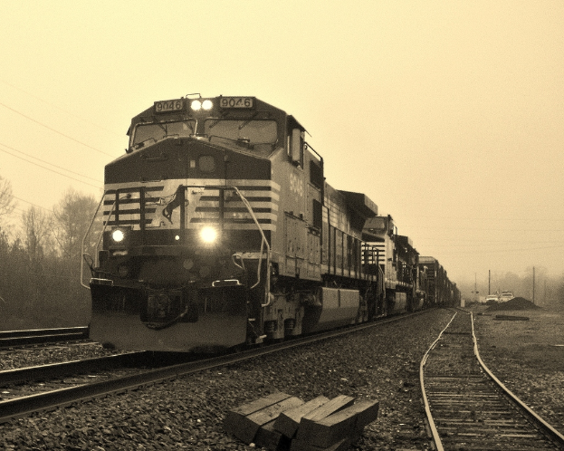 Ein Zug auf den Eisenbahnschienen mit Autos, Strommasten, Bäumen und einem Himmel im Hintergrund.