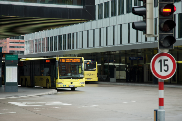 Zwei gelbe Busse fahren auf einer von hohen Gebäuden gesäumten Straße, mit einer Ampel und Passanten im Hintergrund.