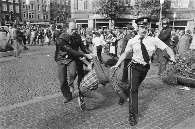 Schwarz-weißes Bild eines Mannes, der von zwei Polizisten auf einer Straße mit Zuschauern, Bäumen, Gebäuden, Laternen, Verkehrszeichen, Topfpflanzen und einem Mann im Vordergrund mit Schuhen gezogen wird.