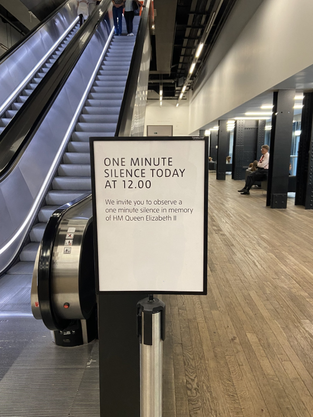 Eine Rolltreppe im Flughafen mit einem Schild, auf dem "Eine Minute Stille heute" steht, einige Menschen darauf und an der Decke angebrachte Lampen im Hintergrund.