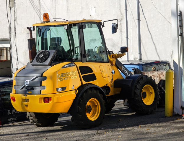 Ein gelber Radlader auf einer Straße vor einem Gebäude mit einer Wand im Hintergrund, umgeben von anderen Fahrzeugen.