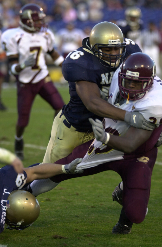 Zwei Fußballspieler in Helmen und Uniformen tackeln sich auf dem Feld, während andere im Hintergrund zuschauen.