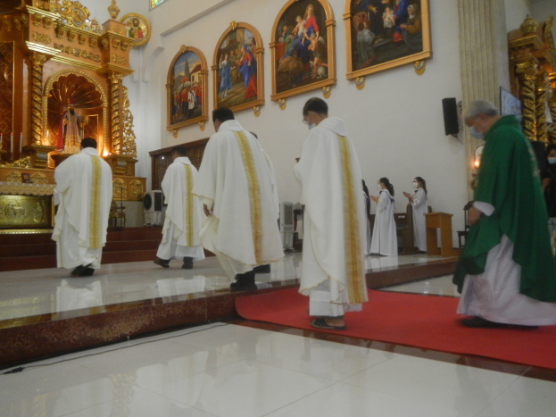 Eine Gruppe von Priestern geht eine rote Teppich in einer Kirche entlang, mit Menschen, die auf dem Boden vor und rechts von ihnen stehen, und Fotorahmen an der Hintergrundwand.