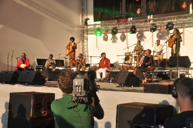 Ein musikalisches Konzert mit Musikern, die verschiedene Instrumente auf der Bühne spielen, darunter Gitarren und Trommeln, unter rot und grünen Scheinwerfern, mit Lautsprechern unten und zwei Personen, eine Aufnahme und die andere zuhört.