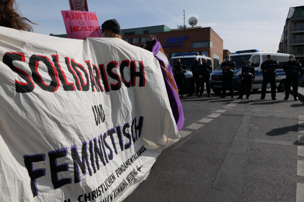 Gruppe von Menschen marschiert auf einer Straße und hält ein 'Solidarität und Feminismus'-Schild, mit parkenden Fahrzeugen und Gebäuden im Hintergrund unter einem klaren blauen Himmel.