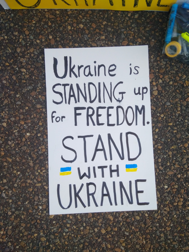 Ein weißer Papierbogen mit den Worten "Ukraine steht für Freiheit auf, stellt Euch hinter Ukraine" liegt vor einem gelben Banner auf dem Boden. Rechts davon befindet sich eine Plastikhülle mit verschiedenen Gegenständen.