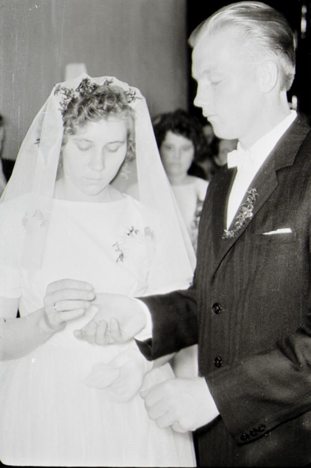 Ein Schwarz-Weiß-Foto einer Braut in einem weißen Kleid und Schleier und eines Bräutigams in einem Anzug, die ihre Eheringe austauschen, mit ein paar Leuten im Hintergrund.