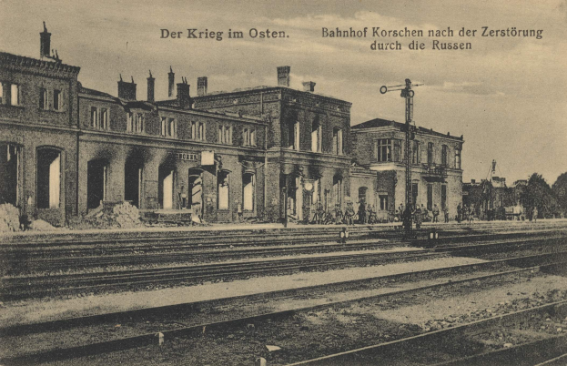 Altes Schwarz-Weiß-Foto des Bahnhofs Korschen in Ostern, Deutschland, zeigt einen Bahnhof mit Bahnschienen, Gebäuden, Pfählen, Menschen, Bäumen und einem Himmel im Hintergrund.