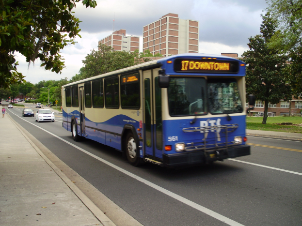 Blauer und weißer Shuttlebus fährt auf einer Straße mit hohen Gebäuden, Menschen auf dem Bürgersteig, Bäumen, Polen und einem klaren blauen Himmel im Hintergrund.