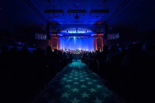 Ein großer Saal mit sitzenden Zuschauern, eine Bühne mit Musikern und Deckenleuchten und -fernseher, was auf eine Konzerthalle hinweist.