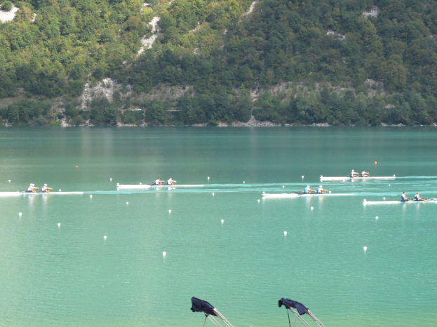 Eine Gruppe von Menschen, die in einem Wettbewerb auf einem Gewässer mit Bäumen und einem Hügel im Hintergrund rudern, mit Kleidung, die über Stände am Boden gehängt ist.
