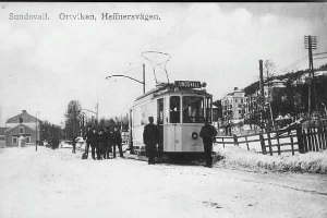 Ein Schwarz-Weiß-Foto einer Tram auf einer verschneiten Straße mit Menschen daneben, ein Zaun auf der rechten Seite und im Hintergrund Gebäude, Bäume, Pfosten, Drähte und der Himmel, mit Text oben.