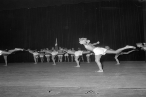 Schwarzes Foto einer Gruppe, die auf einer Bühne Gymnastik macht, mit einem Vorhang und einer Fahne im Hintergrund.