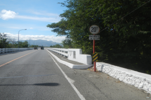 Eine Fahrbahn mit Fahrzeugen, ein Tempolimit-Schild, Geländer, Laternen, Bäume und ein bewölkter Himmel.