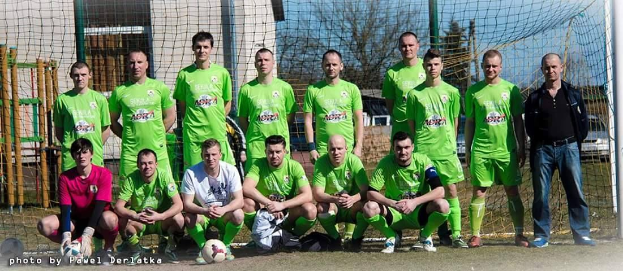 Männergruppe in grünen T-Shirts auf einem Fußballfeld neben einem Torpfosten stehend, mit einem Ball auf dem Boden.